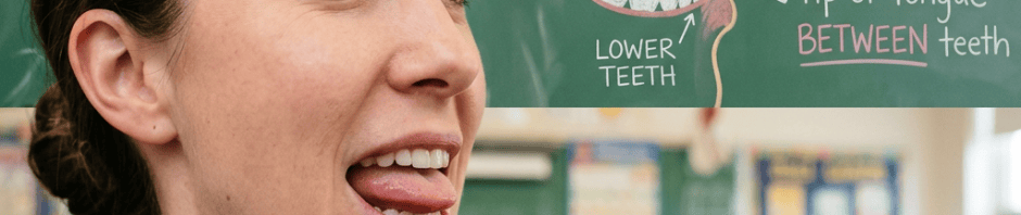 Teacher demonstrating “th” sound; chalkboard reads “THE /th/ SOUND,” “TONGUE TIP,” and “BETWEEN teeth.”